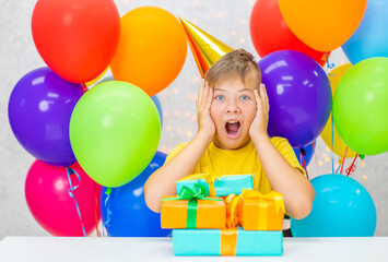 Shocked young boy wearing party's cap sits with many gift boxes. Empty space for text