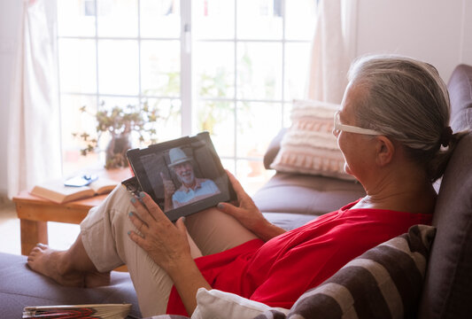 Senior Woman Red Dressed Lying In Sofa At Home Using Tablet Device To Video Call With Husband Absent Due To Coronavirus - On The Screen Smiling Bearded Man Doing Ok Sign With Hand