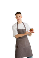 Whiskey. Portrait of a young male caucasian barista or bartender in brown apron smiling. Studio white background, copyspace. Holding cocktails, inviting guests. Professional occupation, drink, service