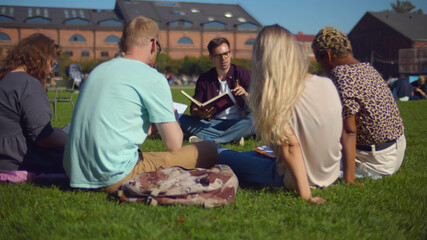 Group of college friends preparing for lessons outdoors, sitting on lawn in park, studying together