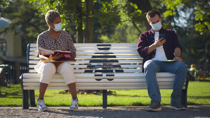 Two students wearing masks sitting on bench keeping social distance and studying
