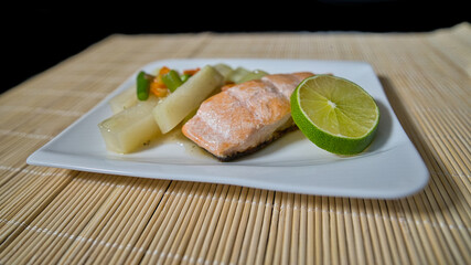 Gebratener Lachs mit Kohlrabi Gemüse, Karotten, Bohnen und einer Scheibe Limette auf einem weißen Teller serviert
