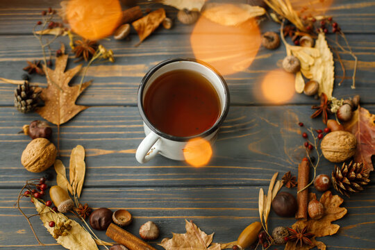 Vintage metal mug with tea and autumn wreath made of leaves, berries, acorns and pine cones on dark wooden background with warm lights. Hello fall! Rustic autumn image