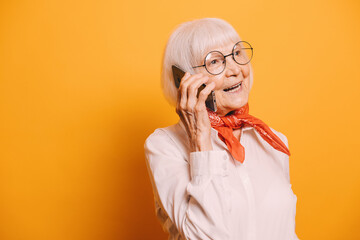 Image of senior gray-haired woman with gray eyes wearing white blouse and orange scarf or cravat standing on isolated over orange background, looking sideway, smiling and talking by her smartphone.