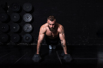 Young strong sweaty fit muscular man with big muscles doing push ups with dumbbell weights on the...
