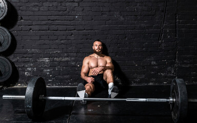 Young strong fit sweaty muscular man sitting on the floor of the gym after barbell weight lifting workout and taking a break from hardcore cross training