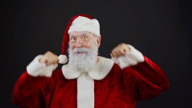 Medium shot portrait of joyful Santa Claus in traditional red costume and eyeglasses looking at camera while dancing funny dance against black studio background