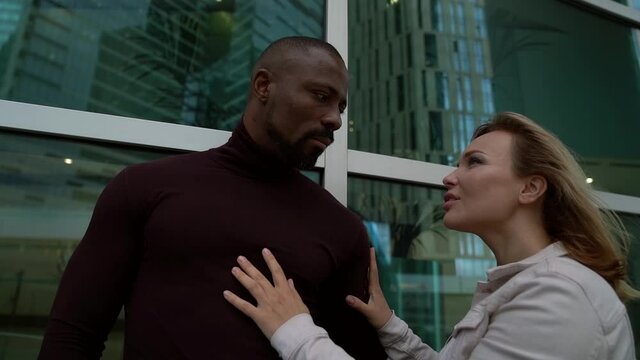 A African-American man and a white woman look at each other with loving eyes against the background of a business center window. Beautiful interracial couple are on a city street.