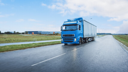 Long Haul Semi-Truck with Cargo Trailer Full of Goods Travels on the Highway Road. Daytime Driving Across Continent Through Rain. Industrial Retail Warehouses Area. Front Shot