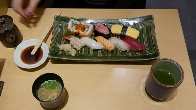 Woman tip soy sauce to saucer and split chopsticks, top down view of table. Rectangular platter with assorted sushi, miso soup cup and traditional green tea on table