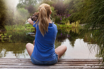 Pretty woman with long hair sitting in zen garden in front of lake and holding cute dog. Calming power of dogs.