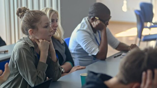 Young multi-ethnic students are having study time at table in university room irrl. Multiracial guys and girls listening lecture and sitting at desk, african american man using gadget. Diverse