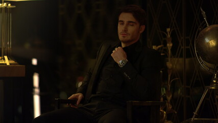 Obraz na płótnie Canvas Hot guy touching shirt collar indoors. Handsome boss sitting armchair dark room