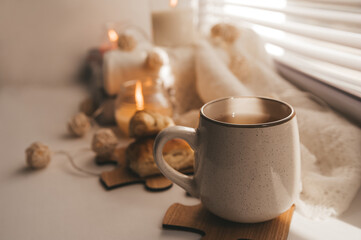 A cozy and warm hygge concept with white sweaters, candles, a cup, a garland on the windowsill. Autumn trends