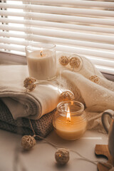 Fototapeta na wymiar A cozy and warm hygge concept with white sweaters, candles, a cup, a garland on the windowsill. Autumn trends