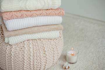 Fototapeta na wymiar A stack of knitted sweaters, cotton, a candle. Women's blouses are on the ottoman. Cozy autumn clothes.