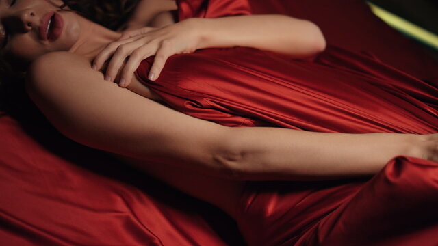 Sexy woman hands playing red silk sheet in bed. Naked girl relaxing in bedroom.