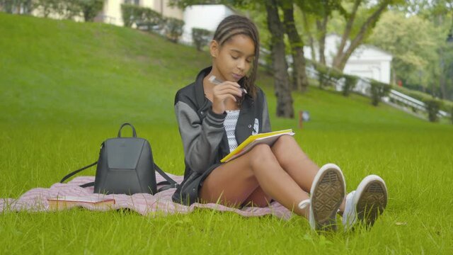Pretty African American schoolgirl doing homework on sunny green lawn in park. Portrait of intelligent little girl writing in workbook and thinking. Education and intelligence.
