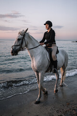 white horse with a woman riding stands on the beach in the evening