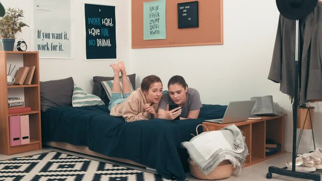 Dolly-in shot of young female friends lying together on bed in cozy room and chatting while looking at mobile phone