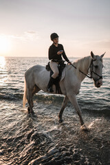 Fototapeta premium young woman with phone rides astride a white beautiful horse on the beach at sunset