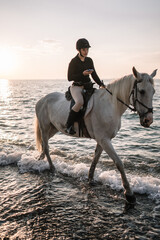 young woman with a phone rides a white beautiful horse and communicates in social networks on the beach at sunset