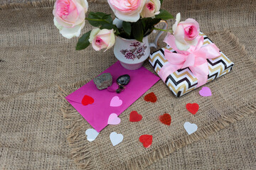 There is a bouquet of roses in a vase on the table. Nearby lies a gift box, a pink envelope, earrings and two natural semi-precious stones, black onyx and arsenopyrite..