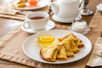 a few pancakes on a plate, honey in a gravy boat. In the background there is a set table, on the table there is a teapot, a cup of tea. Breakfast with pancakes.
