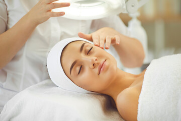 Obraz na płótnie Canvas Smiling young womans face under light getting and enjoying facial massage from cosmetologist