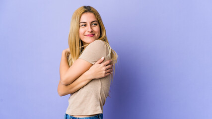 Young blonde woman isolated on purple background hugs, smiling carefree and happy.