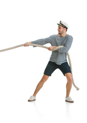 Pulls the rope, tug of war. Caucasian male sailor in uniform on white studio background. Young man using modern devices and gadgets. Concept of tech, professional occupation, job of seaman, emotions.