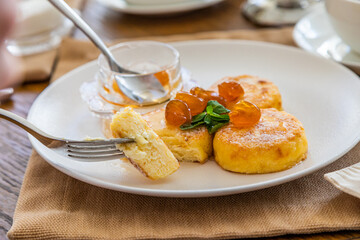 The curd cakes covered in jam are cut with a knife and pricked on a fork. breakfast with cheesecakes with jam. In the background a cup with tea.