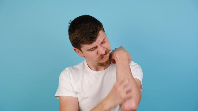 Unhappy tired young man scratching her itchy arm, dressed in white t-shirt, isolated on blue studio background for advertisement. Itch symptoms from insect bite or chemical allergy cosmetic