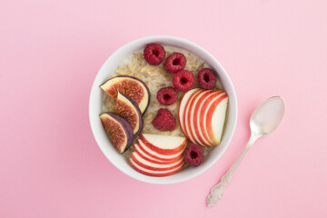 Top view of milk oatmeal with figs, raspberries and apple  in the white bowl in the center of the pink background