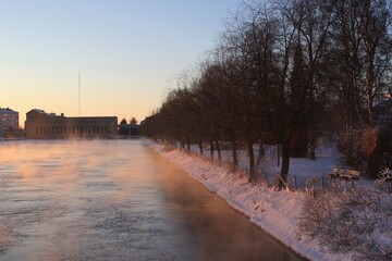 A sunset by a dammed river on a very cold winter day. The steam and snow is colored pink by the sun. Behind, there are urban buildings.