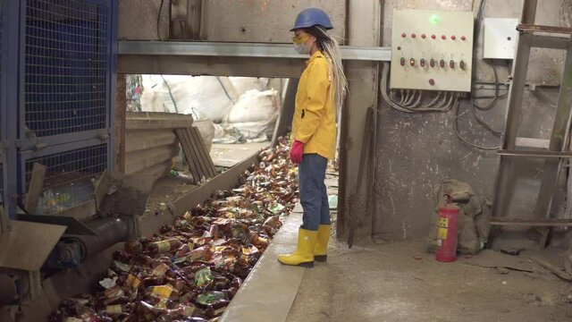 Woman-worker in yellow and transparent protecting glasses, hard hat and mask watching the conveyor full of used plastic bottles. Footage of automized process on recycle plant