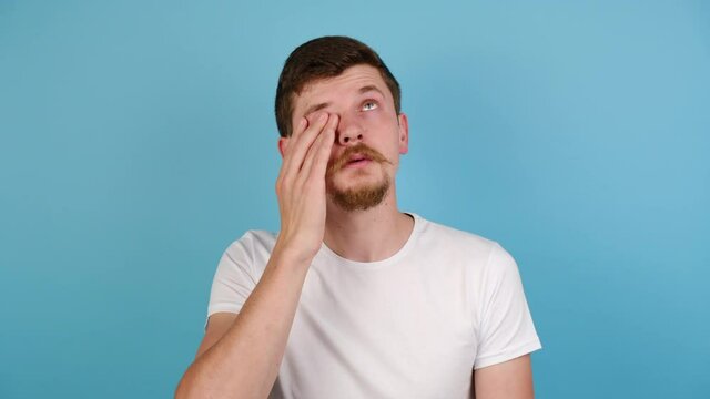 Tired young man massaging nose bridge, feeling eye strain, exhausted guy suffering from headache or migraine, wears t-shirt, isolated on blue studio background with copy space. Eyes fatigue concept