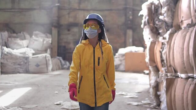 Waste processing plant. Recycling and storage of waste for further disposal. Woman worker in hard hat, gloves and mask walking through the stacks of pressed disposal waste, huge piles. Front view
