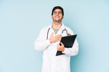 Young doctor man isolated on blue background laughs out loudly keeping hand on chest.