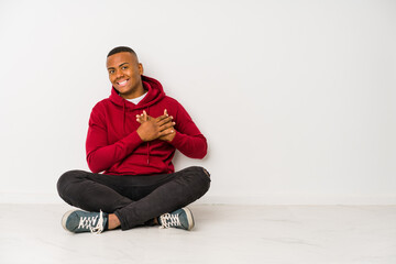 Young latin man sitting on the floor isolated has friendly expression, pressing palm to chest. Love concept.