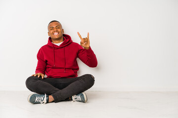 Young latin man sitting on the floor isolated showing a horns gesture as a revolution concept.
