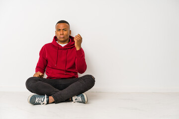 Young latin man sitting on the floor isolated showing fist to camera, aggressive facial expression.