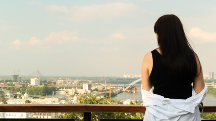 Fototapeta na wymiar Back view of a young woman in a black T-shirt and white shirt, a brunette traveler looks at the cityscape on a summer sunny day from a high hill with an observation deck for tourists. Vintage