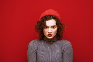 Closeup portrait of a curly-haired lady in a gray sweater with a hat on her head looking into the camera with a serious face. Serious young model in hat and freckles isolvoan on red background.