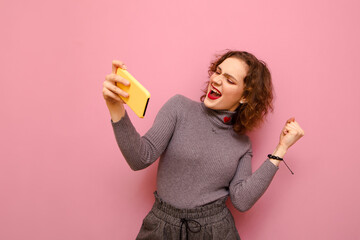 Pretty lady with a happy face and smartphone in hand is pleased to win games. Curly attractive lady in gray sweater is happy, emotionally celebrates victory with a smartphone in her hand.