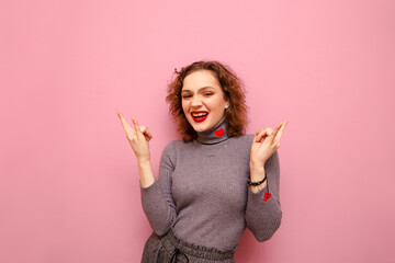 Joyful girl with curly hair and gray sweater stands on a pink background, looks in camera with a...