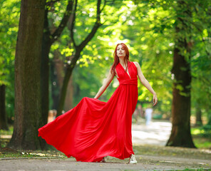 Young fashionable woman in long red luxurious dress walks in summer park