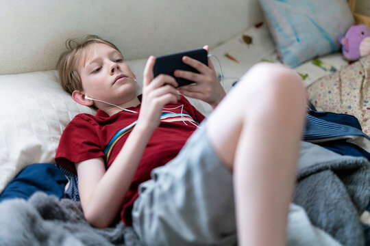 Young boy lying on bed looking at smartphone with earbuds in ears