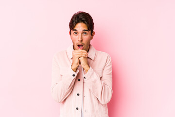 Young caucasian man posing in a pink background isolated praying for luck, amazed and opening mouth looking to front.