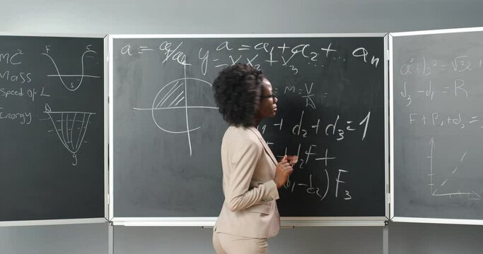 Rear of African American young female teacher of math or physics writing formulas on blackboard with chalk. School tutor woman turning to camera and explaining mathematics. Back view.